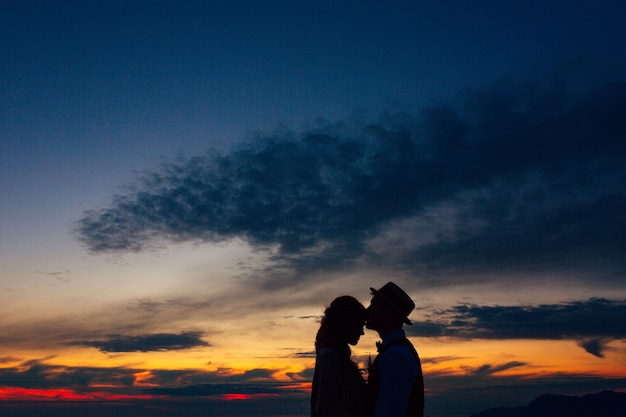 Siluetta dello sposo con il cappello bacia la sposa sulla fronte al tramonto