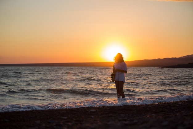 Siluetta di giovane donna sexy che gioca sassofono sulla spiaggia al tramonto