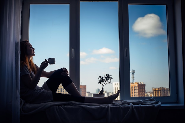 Siluetta di giovane ragazza snella che si siede sul davanzale della finestra a casa, vista laterale, spazio della copia. Fuori dalla finestra il cielo azzurro e gli edifici alti della città. Donna dai capelli rossi che tiene tazza, guardando la metropoli.