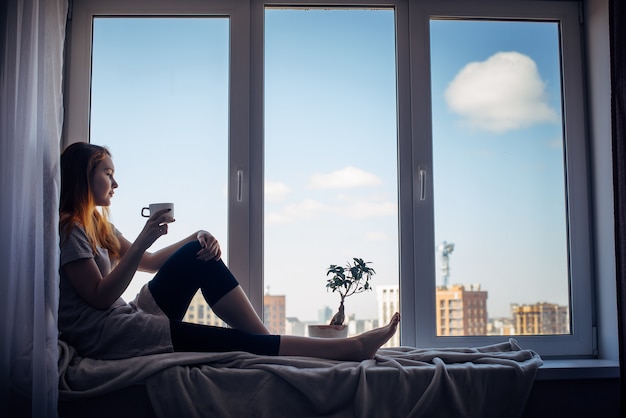 Siluetta di giovane ragazza snella che si siede sul davanzale della finestra a casa, vista laterale, spazio della copia. Fuori dalla finestra il cielo azzurro e gli edifici alti della città. Donna dai capelli rossi che tiene tazza, guardando la metropoli.