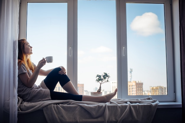 Siluetta di giovane ragazza snella che si siede sul davanzale della finestra a casa, vista laterale, spazio della copia. Fuori dalla finestra il cielo azzurro e gli edifici alti della città. Donna dai capelli rossi che tiene tazza, guardando la metropoli.