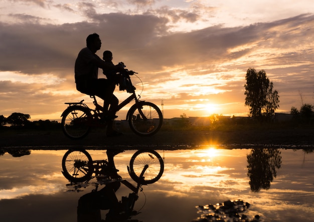 Siluetta di riflessione del padre con il suo bambino in bicicletta contro il tramonto.