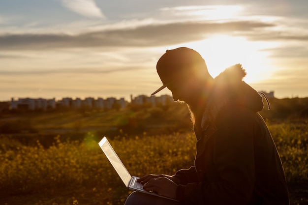 Siluetta di un giovane con una barba che lavora con il suo computer portatile al tramonto in natura