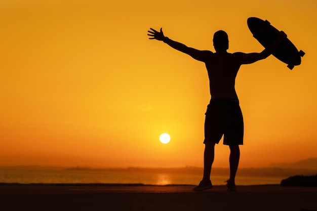 Siluetta di un uomo libero che tiene uno skateboard al tramonto luminoso