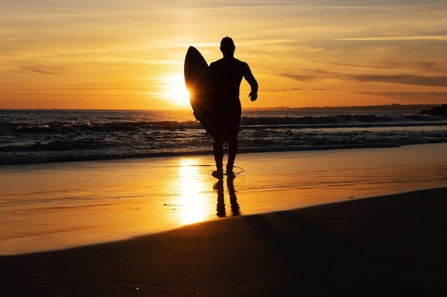 Siluetta di un uomo surfista che cammina in riva al mare al tramonto