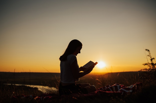 Siluetta di una giovane bella donna al tramonto che si siede su un plaid e fissando attentamente il libro aperto.