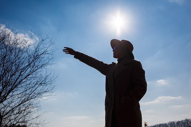 Siluetta di una ragazza con cappello e cappotto su uno sfondo di cielo con nuvole e sole.