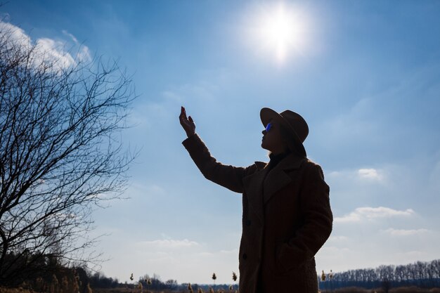 Siluetta di una ragazza con cappello e cappotto su uno sfondo di cielo con nuvole e sole.