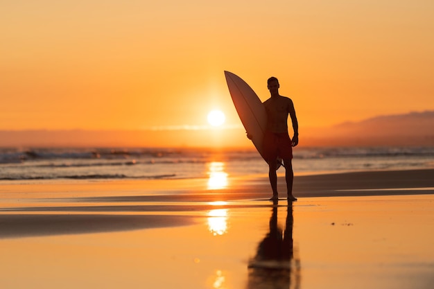 Siluetta nera di un uomo attraente sulla riva che tiene una tavola da surf al tramonto arancione