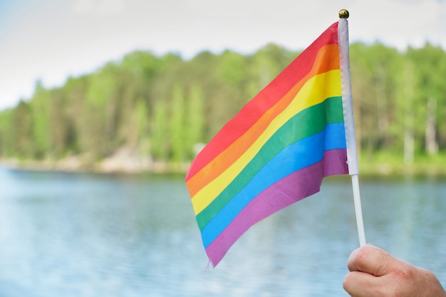 Simbolo della bandiera arcobaleno della comunità lgbt su uno sfondo di erba, lago e alberi, bandiera in mano di donna