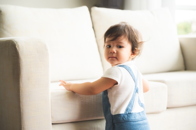 Simpatica bambina giocosa che impara a camminare e stare in piedi sostenendosi al divano di casa mentre distoglie lo sguardo maliziosamente