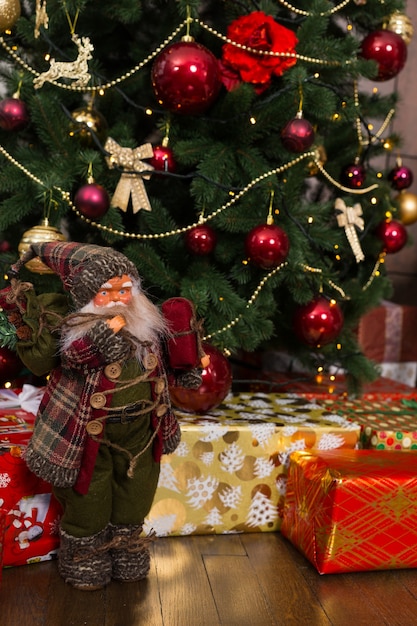Simpatica bambola di Babbo Natale con un rossore sulle guance che indossa un costume lavorato a maglia e tiene un regalo in piedi vicino all'albero di Natale con regali