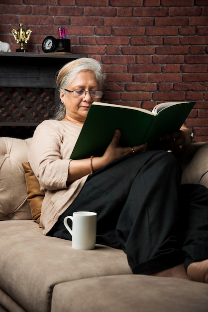 Simpatica signora asiatica indiana anziana seduta su una sedia reclinabile o su un divano leggendo un libro o usando una scheda o un computer portatile