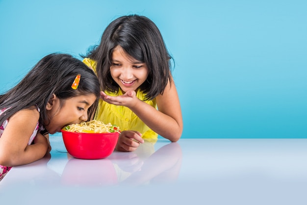 Simpatiche bambine indiane o asiatiche che mangiano gustose tagliatelle cinesi con forchetta o bacchette, isolate su sfondo colorato colorful