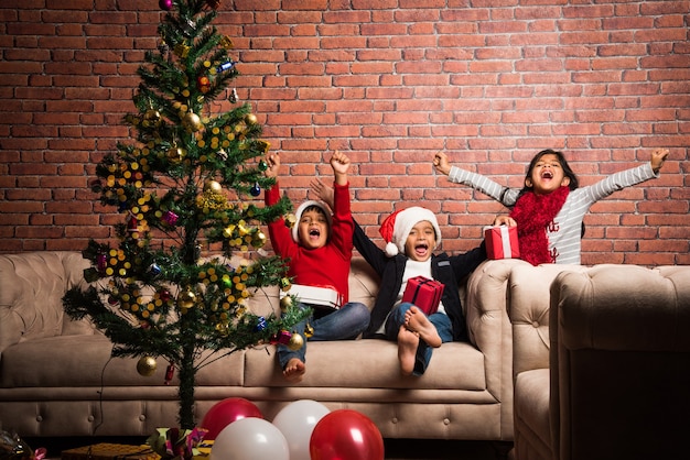 Simpatici bambini asiatici indiani che festeggiano il Natale a casa con cappello da Babbo Natale, regali e albero di Natale