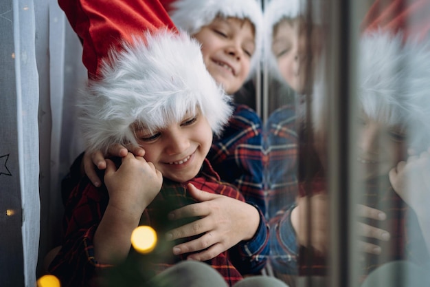 Simpatici bambini caucasici che indossano cappelli di snta e camicie a quadri seduti abbracciati vicino alla finestra in attesa di Natale