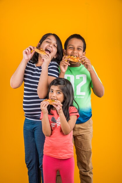 Simpatici bambini indiani o asiatici che mangiano gustosi hamburger, panini o pizza in un piatto o in una scatola. In piedi isolato su sfondo blu o giallo.