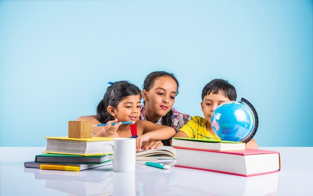 Simpatici bambini indiani o asiatici che studiano sul tavolo da studio con una pila di libri, globo educativo, isolato su colore azzurro