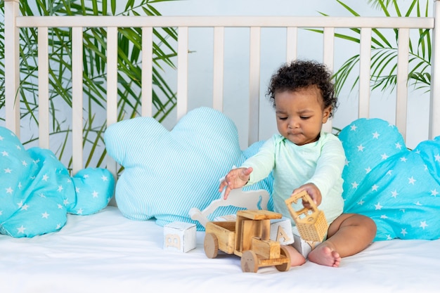 Simpatico bambino africano seduto a letto a casa su un letto di cotone blu e giocando con i giocattoli di legno.