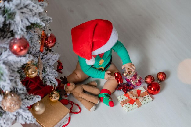 Simpatico bambino con un body verde e un cappello da Babbo Natale decora l'albero di Natale con palloncini colorati. bambino con un costume da gnomo di Natale. concetto di Capodanno invernale. spazio per il testo. vista dall'alto