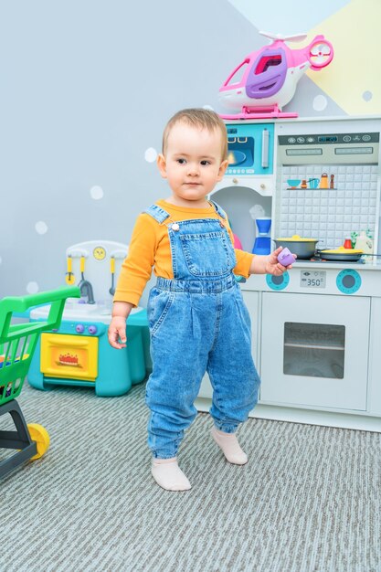 Simpatico bambino di 23 anni nella sala giochi vestito con una tuta di jeans che gioca con una tazza di plastica nella cucina dei giocattoli