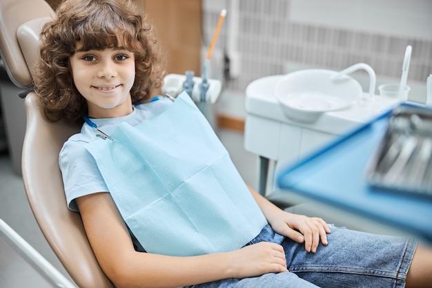 Simpatico bambino riccio che sorride mentre aspetta un dentista in una clinica odontoiatrica