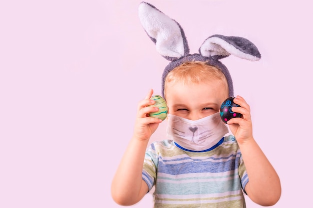 Simpatico neonato con orecchie da coniglio sulla testa e maschera protettiva con uova colorate su sfondo rosa. Bambino felice sorridente allegro. Banner per le vacanze di Pasqua di Covid