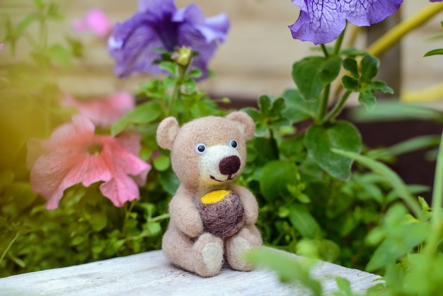 Simpatico orsetto giocattolo fatto a mano si siede con un barile di miele tra i fiori, feltro lana