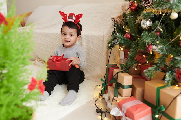 Simpatico ragazzino che indossa una fascia con corna di renna quando è seduto accanto a un albero di Natale decorato con un regalo in mano