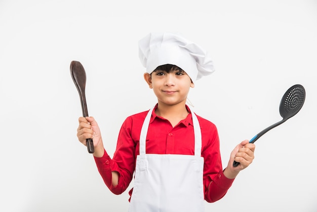 Simpatico ragazzino indiano o ragazzo vestito da chef e con in mano utensili da cucina, in piedi isolato su sfondo bianco