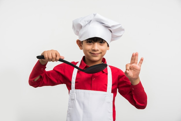 Simpatico ragazzino indiano o ragazzo vestito da chef e con in mano utensili da cucina, in piedi isolato su sfondo bianco