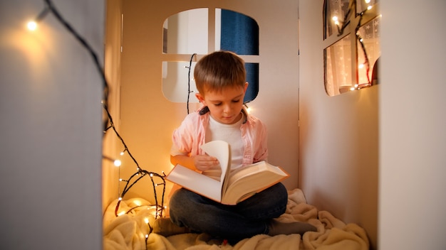 Simpatico ragazzino seduto in una casa di cartone giocattolo di notte e leggendo un grande libro di fiabe. Concetto di educazione dei bambini e lettura in camera oscura.