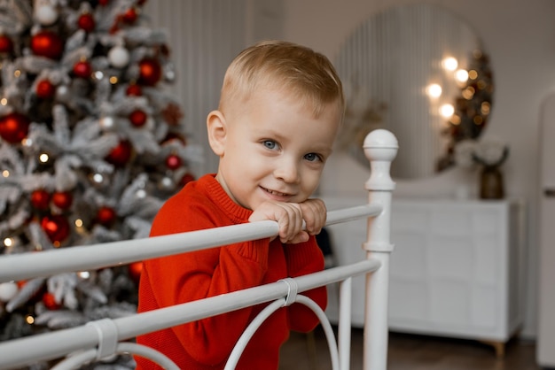 Simpatico ragazzino sorridente con un maglione rosso seduto sul letto sullo sfondo dell'albero di Natale