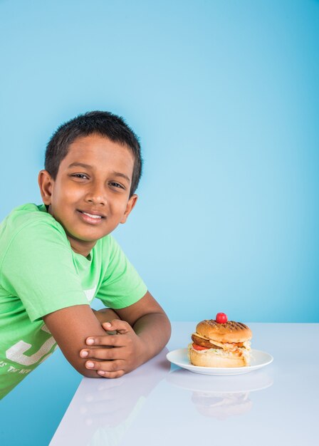 Simpatico ragazzo indiano che mangia hamburger, piccolo ragazzo asiatico e hamburger, su sfondo blu