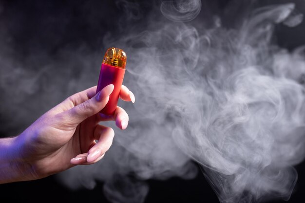 Sistema di vape pod di colore rosso con cartucce sostituibili in mani femminili isolate su sfondo nero Il concetto di moderno svapo di fumo e nicotina sullo sfondo del fumo