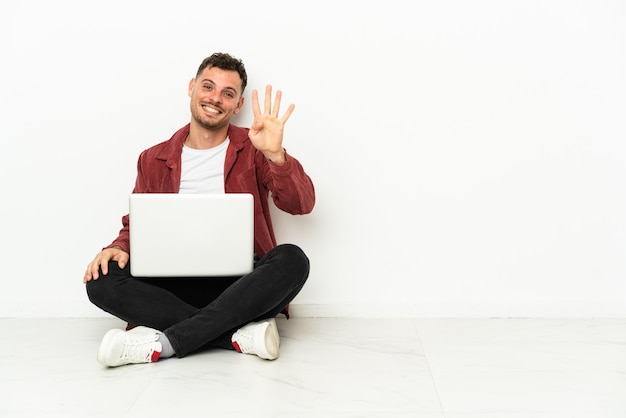 Sit-in di giovane uomo caucasico bello sul pavimento con il computer portatile felice e contando quattro con le dita