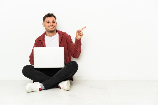 Sit-in di giovane uomo caucasico bello sul pavimento con il dito puntato del computer portatile al lato