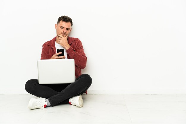 Sit-in di giovane uomo caucasico bello sul pavimento con il pensiero del computer portatile e l'invio di un messaggio