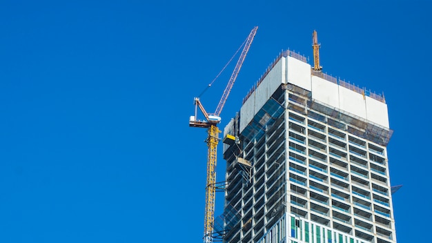 Sito della costruzione di edifici e della gru contro cielo blu con il tabellone per le affissioni bianco in bianco per la pubblicità alla cima di architettura della torre.