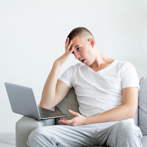 Situazione senza indizi Uomo confuso Conferenza online Dispiaciuto scioccato casual ragazzo seduto sul divano guardando laptop facepalm mano sulla testa nell'interno della stanza luminosa