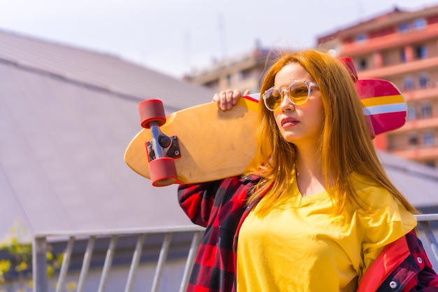 Skateboarder donna in una camicia gialla, camicia a quadri rossa e occhiali da sole, in posa con lo skateboard rivolto a sinistra