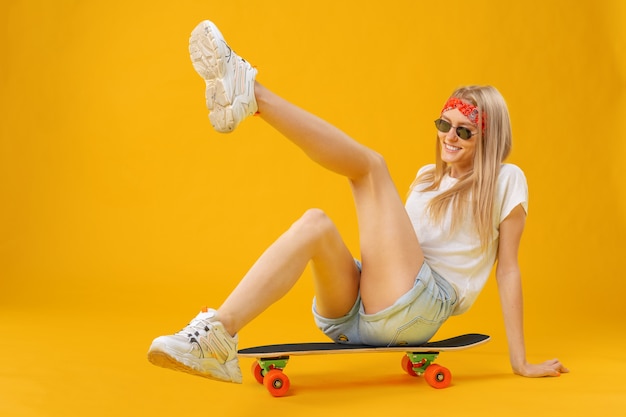 Skateborder ragazza in pantaloncini e maglietta seduto a bordo su giallo