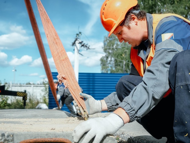 Slinger posa lastra di cemento sul cantiere il giorno d'estate Lavoratore in giubbotto protettivo e casco da costruzione supervisiona la posa della base sul cantiere