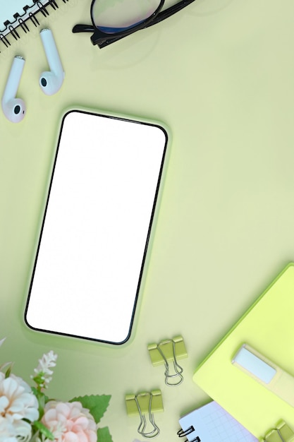 smart phone con schermo vuoto, auricolari, fiori e notebook su sfondo verde.
