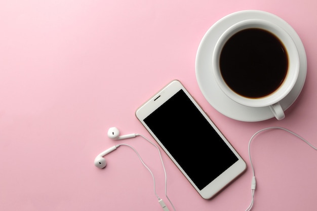 Smartphone bianco con cuffie e una tazza di caffè su uno sfondo rosa brillante. vista dall'alto