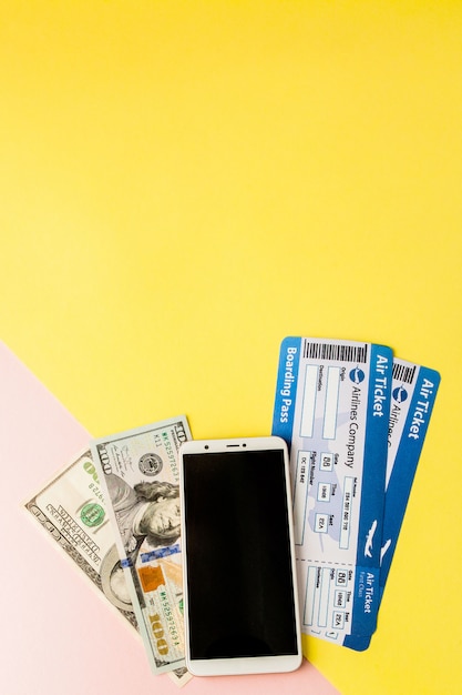 Smartphone, biglietto aereo e dollari su sfondo rosa e giallo pastello. Stile minimal, flatlay.