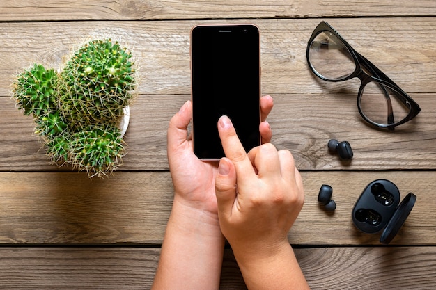 Smartphone con display a schermo nero in mani femminili, cuffie wireless, cactus, capsula di ricarica