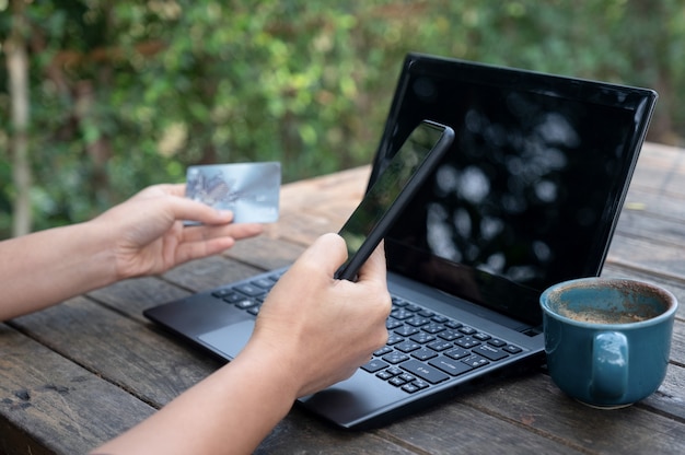 Smartphone e carte di credito in mano con laptop e caffè sul tavolo di legno.