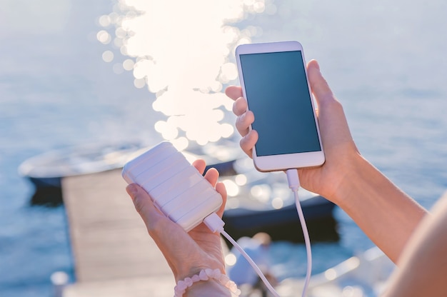 Smartphone in mano a una ragazza sul molo. Ricarica il tuo telefono con Power Bank. Sullo sfondo di un fiume, lago, con un ponte e barche.