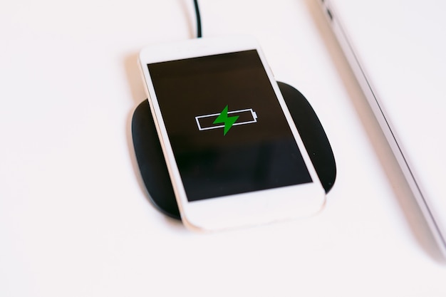 Smartphone mobile bianco con il logo di una batteria con un fulmine verde sullo schermo, in carica su una base di ricarica senza cavo accanto al laptop su un tavolo da lavoro bianco